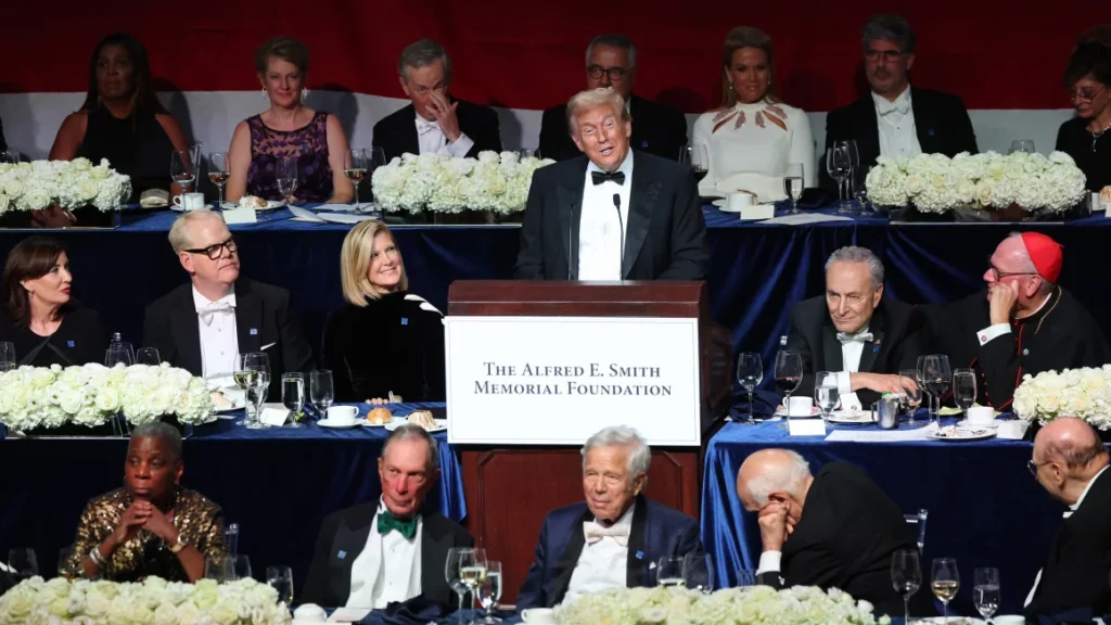 Donald Trump gestures at the podium during the 2024 Al Smith Dinner, with guests in tuxedos and gowns listening intently.